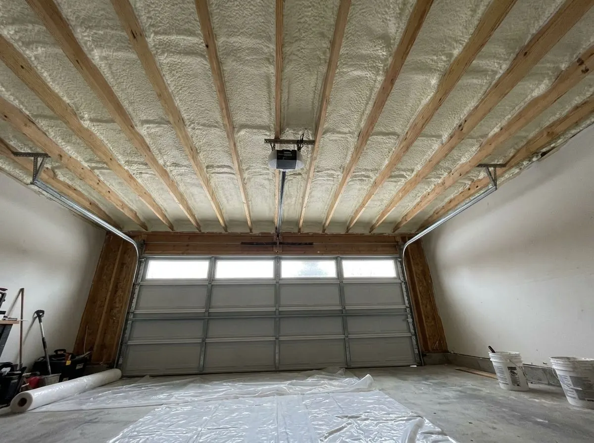 An attached garage with the ceiling drywall removed, showing floor joists above with the underside of the subfloor sprayed with yellow foam.