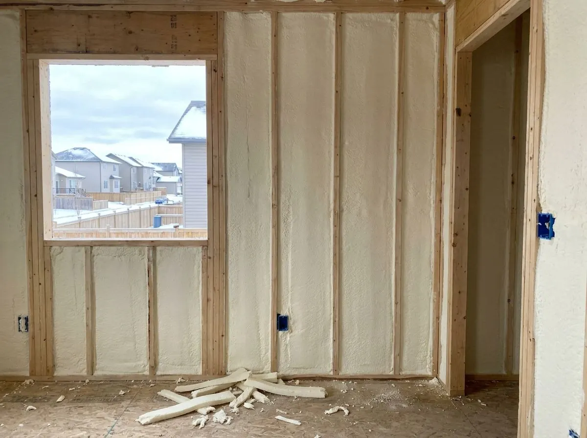 Soft, off-white open-cell spray foam fills the stud cavities of an exterior wall around a window opening, trimmed flush with the studs, with foam offcuts on the subfloor.