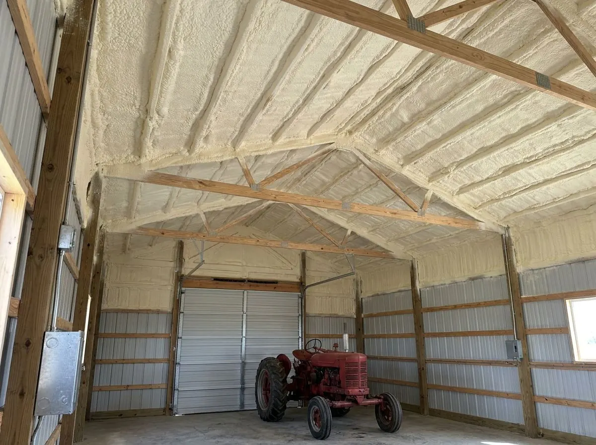 Inside a post-frame shop, the underside of the steel roof and wall panels between wood purlins is covered in yellow spray foam above a tractor.