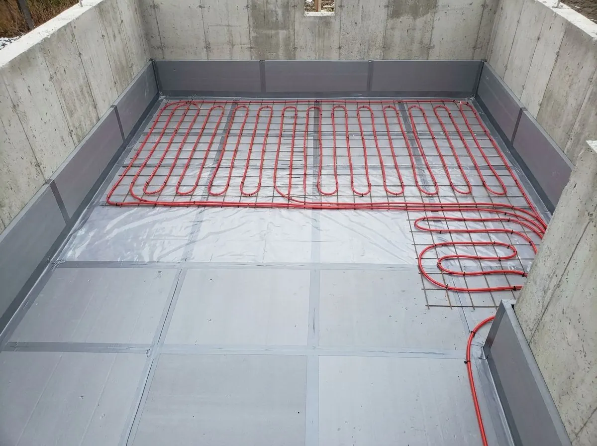A basement floor area covered with rigid foam insulation boards and a sheet of poly, with red PEX radiant heating tubing clipped in loops on top.